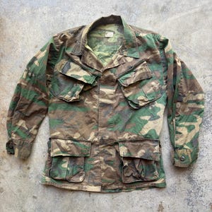 Può includere: Giacca con motivo mimetico con quattro tasche anteriori e due tasche sul petto. La giacca presenta una chiusura con bottoni e maniche lunghe. La tavolozza dei colori comprende sfumature di verde, marrone e beige, creando un'estetica di ispirazione militare.