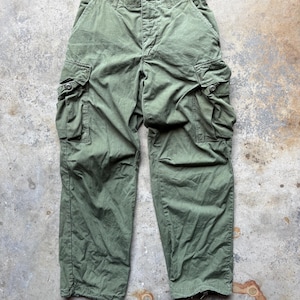 Könnte beinhalten: Olivgrüne Cargohose mit mehreren Taschen und Knopfverschlüssen. Die Hose hat eine lockere Passform und ein abgenutztes Aussehen, was darauf hindeutet, dass sie Vintage oder gebraucht ist. Der Stoff scheint eine strapazierfähige Baumwollmischung zu sein.