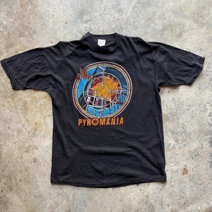 Könnte beinhalten: Schwarzes T-Shirt mit einem kreisförmigen Grafikdesign, das ein Ziel darstellt, und dem Wort "PYROMANIA" in Orange. Die Grafik enthält blaue, orangefarbene und weiße Elemente. Das T-Shirt liegt flach auf einer Betonoberfläche.