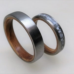 May include: Two wedding bands, one with a silver metal band and a wood inlay, and the other with a silver metal band and a white inlay.