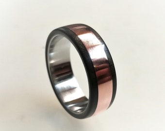 May include: A close-up of a ring with a silver interior, a copper band, and a black outer edge. The ring is made of metal and wood. The ring is a band style and is set against a white background.