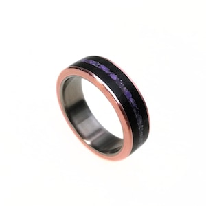 Puede incluir: Un anillo con una banda de color cobre y una incrustación central negra. La incrustación presenta una fina franja de piedras moradas. El anillo tiene un interior de color plateado.