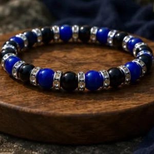 May include: A circular beaded bracelet featuring alternating glossy black and vibrant blue spherical beads. Each bead is separated by a small, silver-toned spacer ring encrusted with tiny, clear, reflective crystals, creating a repeating pattern.