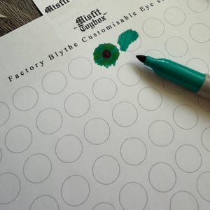 Peut inclure: Gros plan sur une feuille de papier avec le texte "Misfit Toybox Factory Blythe Customisable Eye chips". Le papier comporte plusieurs cercles gris et un marqueur vert. Deux échantillons verts et marron sont en haut à droite.
