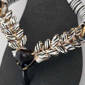 May include: A black flip-flop sandal featuring a thong strap wrapped in white and black striped fabric. The strap is embellished with a row of metallic, leaf-shaped accents and a large, faceted, triangular black stone at the center junction.