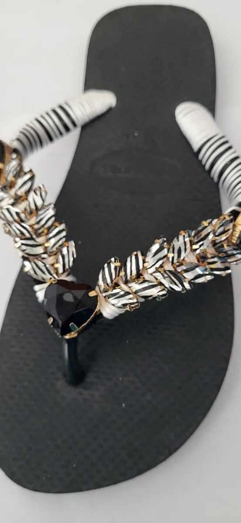 May include: A black flip-flop sandal featuring a thong strap wrapped in white and black striped fabric. The strap is embellished with a row of metallic, leaf-shaped accents and a large, faceted, triangular black stone at the center junction.