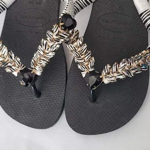 May include: A pair of black flip-flops featuring white wrapped straps adorned with rows of metallic silver and black textured beads. Each strap includes a prominent black heart-shaped gem at the center and a smaller black gem near the toe post.