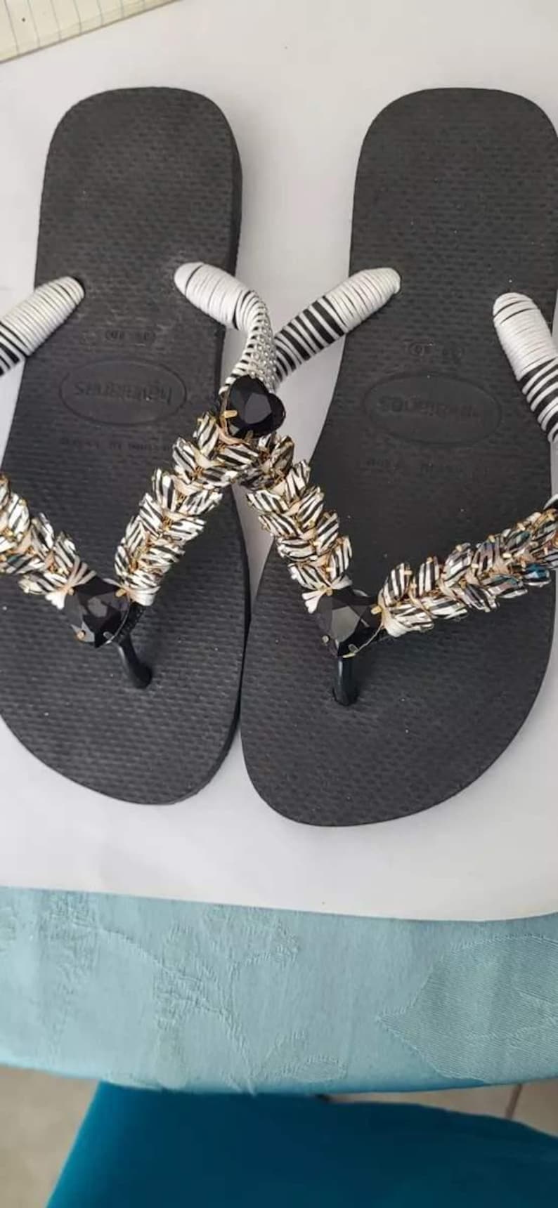 May include: A pair of black flip-flops featuring white wrapped straps adorned with rows of metallic silver and black textured beads. Each strap includes a prominent black heart-shaped gem at the center and a smaller black gem near the toe post.