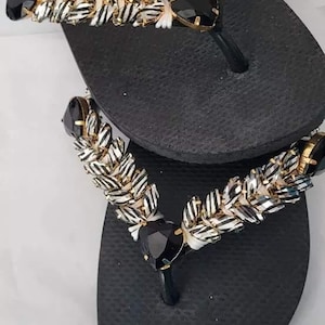 May include: A pair of black flip-flops featuring thong straps embellished with white and black striped fabric, gold-toned metal accents, and large, faceted black heart-shaped stones positioned at the center of each strap assembly.
