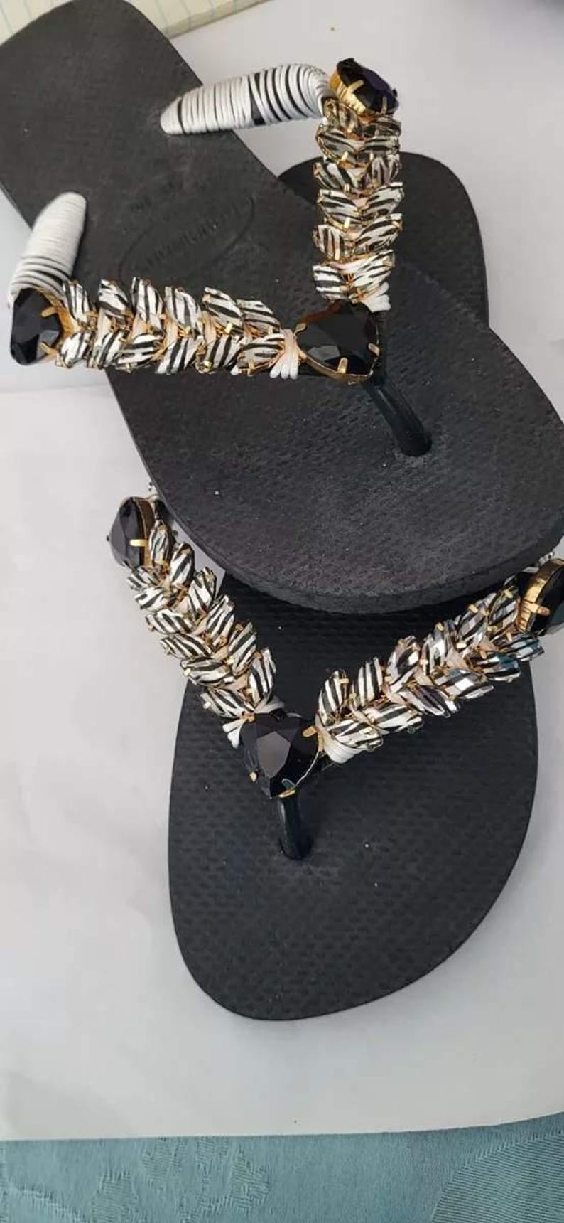 May include: A pair of black flip-flops featuring thong straps embellished with white and black striped fabric, gold-toned metal accents, and large, faceted black heart-shaped stones positioned at the center of each strap assembly.