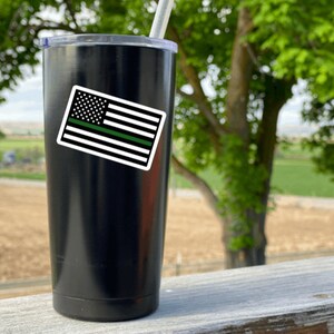 May include: Black stainless steel tumbler with a green line American flag sticker. The tumbler is on a wooden surface with a green field in the background.