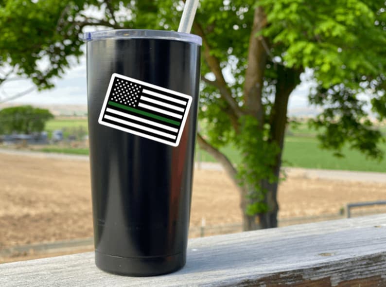 May include: Black stainless steel tumbler with a green line American flag sticker. The tumbler is on a wooden surface with a green field in the background.