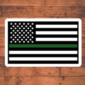 May include: A black and white American flag with a thin green stripe across the middle. The flag is on a wood background.