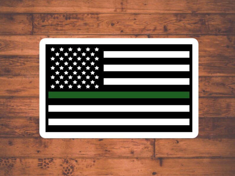 May include: A black and white American flag with a thin green stripe across the middle. The flag is on a wood background.