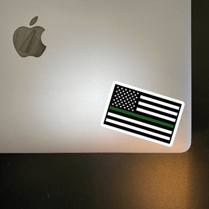 May include: A black and white American flag sticker with a green stripe across the middle. The sticker is on a silver laptop.