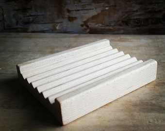 May include: A light-coloured wooden soap dish with a slatted design. The rectangular dish has a series of parallel grooves to allow for drainage. The wood grain is visible, and the dish is set on a wooden surface.