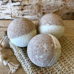 May include: Three spherical bath bombs, each with a two-tone design: a light brown top and a white bottom. They rest on a beige mesh bag with a drawstring closure and a wooden bead. The background is a wooden surface.