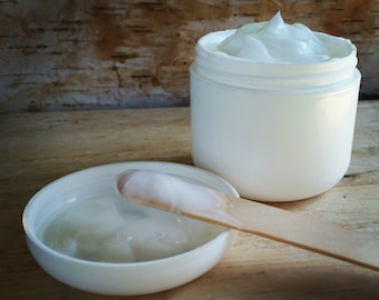 May include: A white jar of cream with a matching lid and a wooden applicator. The jar is open, revealing a thick, white cream. The lid is off, with a small amount of cream inside. The applicator has a dollop of cream on it.