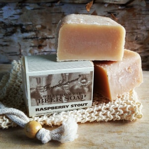 May include: A stack of three light brown beer soaps, one atop a box of soap and a woven washcloth. The box reads "Forever Summer" and "What's on Tap Beer Soap Raspberry Stout". The soap is a natural product.