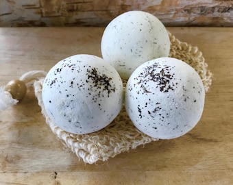 May include: Three white bath bombs with dark speckles rest on a beige, woven washcloth. The bath bombs are spherical and appear to have a textured surface. A wooden bead and a white string are visible on the left side of the washcloth.