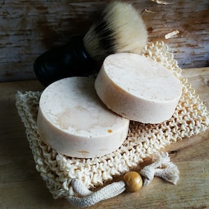 May include: Two round, light-colored soaps rest on a natural fiber soap saver with a wooden bead closure. A black-handled shaving brush with a white and black bristle head is positioned behind the soaps, suggesting a grooming set.
