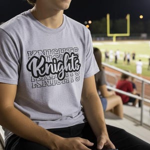 May include: A light grey t-shirt with the word "Knights" in a black, stylised font. The shirt is short-sleeved and appears to be made of a soft material. The background shows a football pitch and spectators.
