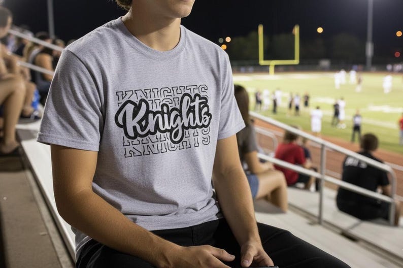 May include: A light grey t-shirt with the word "Knights" in a black, stylised font. The shirt is short-sleeved and appears to be made of a soft material. The background shows a football pitch and spectators.
