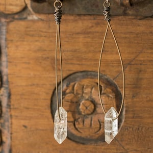 May include: Two pairs of crystal earrings. One pair has a straight wire design, the other a teardrop shape. Both have a clear crystal at the bottom, suspended from a wrapped wire top. The earrings hang against a wooden background.
