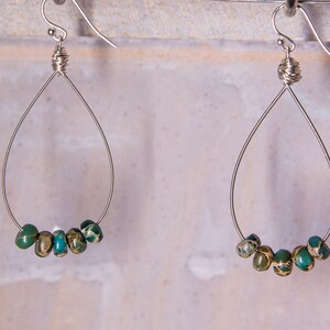 May include: A pair of teardrop earrings. Each earring has a silver wire frame with a cluster of green and teal beads at the bottom. The beads have a speckled pattern. The earrings hang from silver hooks.