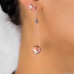 May include: A close-up of a single earring. The earring features a teardrop design with a wire frame and a round, patterned bead in shades of red, brown, and white. The earring is hanging from a silver hook.