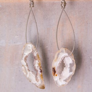 May include: Two agate stone earrings. Each earring features a teardrop-shaped, polished agate stone in white, cream, and brown hues. They are suspended from silver-coloured wire hooks, offering a natural, unique jewellery design.