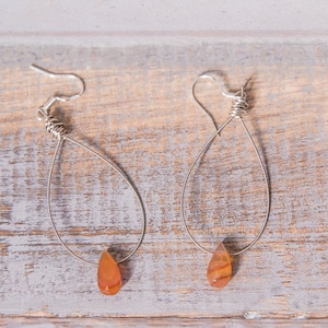 May include: A pair of silver teardrop earrings with orange-brown gemstone drops. The earrings have a simple, elegant design, with the gemstone drops adding a pop of colour. The earrings are displayed on a weathered wooden surface.