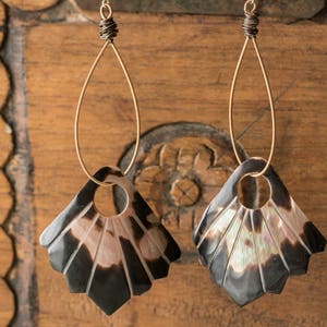 May include: Handmade earrings with a fan-shaped design. The earrings have a black and iridescent shell-like material with gold-coloured wire. They are suspended from a gold-coloured wire and have a circular opening.