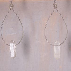 May include: Two silver-coloured wire earrings. Each earring has a teardrop-shaped wire frame with a clear quartz crystal suspended at the bottom. The crystals are elongated and faceted, catching the light. The earrings are displayed against a neutral background.