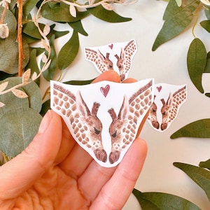 May include: Two watercolor giraffe heads facing each other with a red heart between them. The giraffes are brown and white with a spotted pattern. The sticker is on a white background.