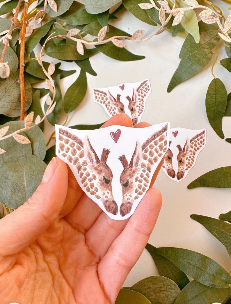 May include: Two watercolor giraffe heads facing each other with a red heart between them. The giraffes are brown and white with a spotted pattern. The sticker is on a white background.