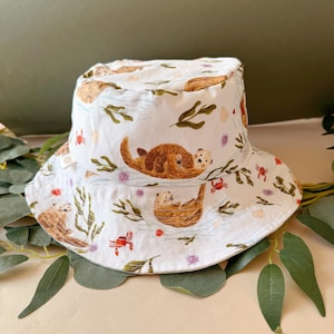 May include: White bucket hat with a repeating pattern of brown otters, green seaweed, red crabs, and purple flowers. The hat has a wide brim and is made of a soft fabric. The hat is designed for sun protection.