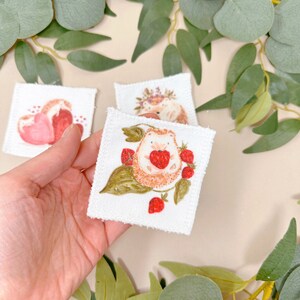 Puede incluir: Un cuadrado de tela blanca con un dise&ntilde;o cosido a mano de un erizo sosteniendo una fresa. El erizo est&aacute; rodeado de hojas verdes y fresas rojas.