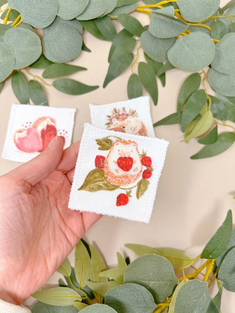 Puede incluir: Un cuadrado de tela blanca con un dise&ntilde;o cosido a mano de un erizo sosteniendo una fresa. El erizo est&aacute; rodeado de hojas verdes y fresas rojas.