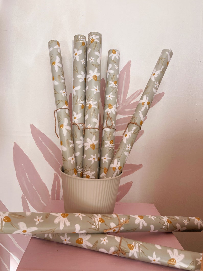Op de afbeelding: Een set van zes rollen groen inpakpapier met een wit bloemenpatroon en gele accenten. De rollen zijn gestapeld in een witte keramische container. Twee extra rollen van hetzelfde inpakpapier liggen op een roze oppervlak.
