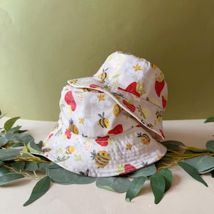 Peut inclure: Deux chapeaux seau blancs avec un motif répété de fraises rouges, d'abeilles jaunes et de fleurs blanches. Les chapeaux sont faits d'un tissu de couleur claire et sont empilés l'un sur l'autre.