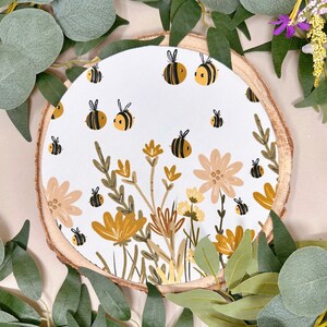 May include: A round wooden slice with a white background featuring a pattern of bees and flowers in shades of yellow, brown, and green.