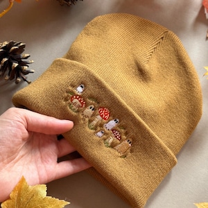 Op de afbeelding: Mosterdgele beanie met een geborduurd paddenstoelontwerp. De omgeslagen rand heeft een cluster van kleurrijke paddenstoelen in rood, wit en bruin. De hoed is gemaakt van een zacht, gebreid materiaal.