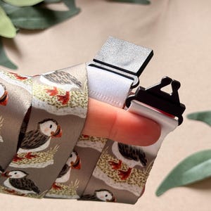 May include: A lanyard with a repeating pattern of puffins on a grey background. The lanyard has a white strap and a black clasp. The puffins are depicted with brown, white, and orange details. The lanyard is designed for holding keys or a badge.