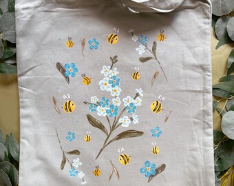 May include: A light gray tote bag with a floral and bee design. The design features blue flowers, yellow and black bees, and brown stems. The tote bag has two handles and is made of a canvas-like material.