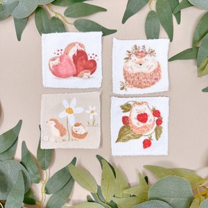 Puede incluir: Cuatro cuadrados de tela blanca con diferentes dise&ntilde;os de erizos. El primer cuadrado muestra una familia de erizos con un coraz&oacute;n rojo. El segundo cuadrado muestra un erizo con una corona de flores. El tercer cuadrado muestra un erizo con una margarita. El cuarto cuadrado muestra un erizo con fresas.