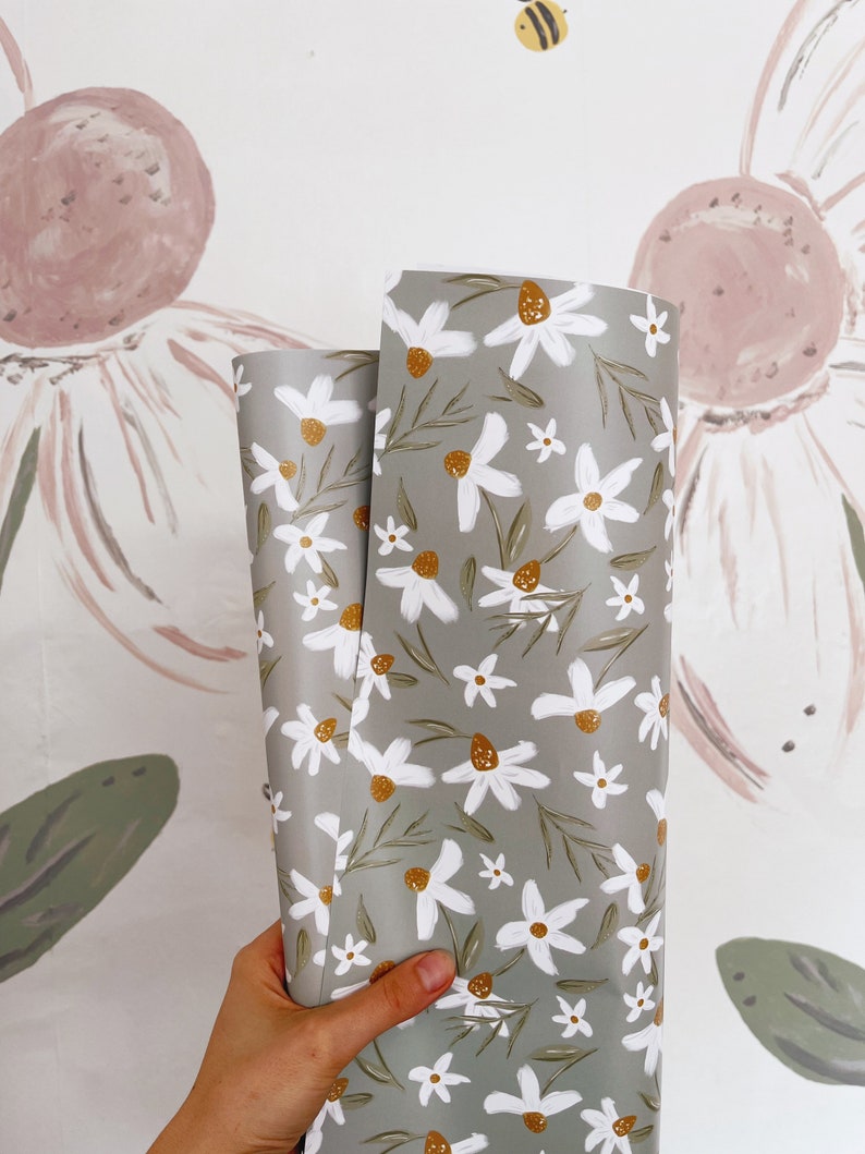 Op de afbeelding: Twee rollen groene bloemenverpakkingspapier met een wit madeliefjespatroon en gouden accenten. Het papier wordt in een hand gehouden.