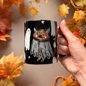 May include: A black ceramic mug featuring a central illustration of a brown bat wearing a grey cape, surrounded by small white ghosts and stars. The design is set against a solid black background, creating a contrast with the whimsical character art.