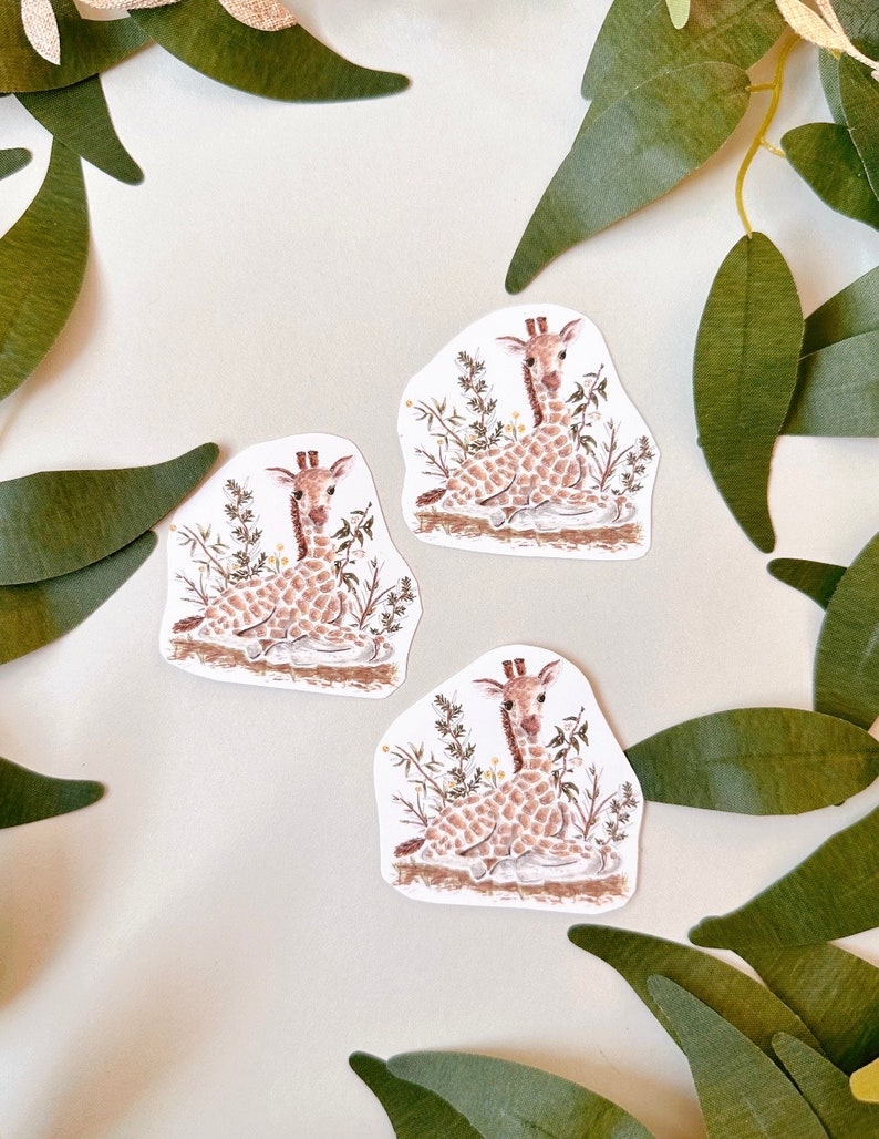 May include: Three stickers featuring a cute cartoon giraffe sitting in a patch of green grass. The giraffe is brown with white spots and has a long neck and long legs. The stickers are perfect for adding a touch of whimsy to your planner, laptop, or water bottle.