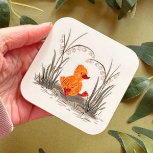 May include: A square coaster featuring a hand-painted illustration of an orange duckling walking through a field of white lily of the valley flowers.